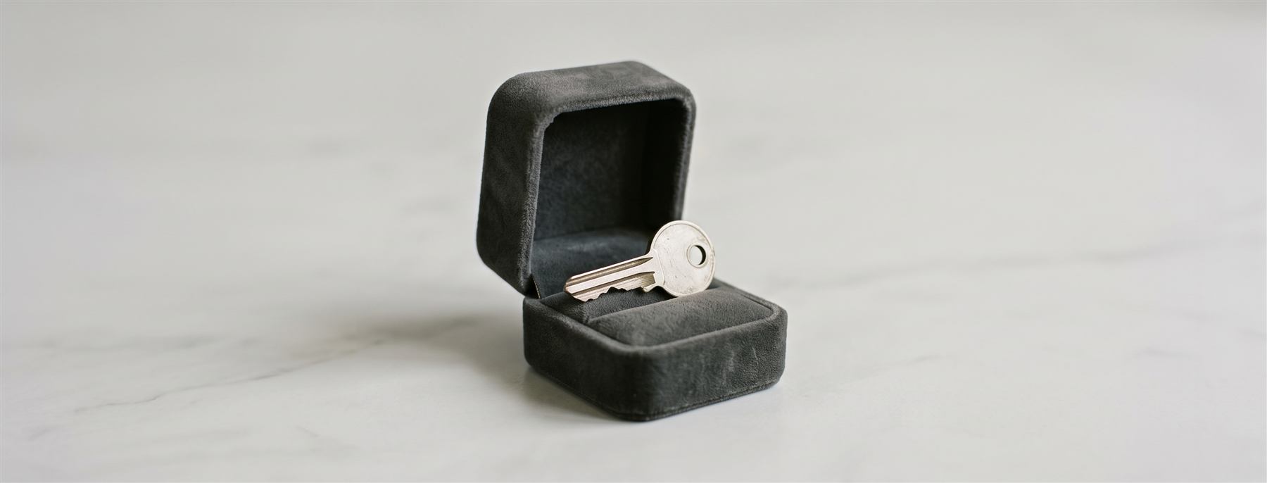 Uma caixa de anel aberta, forrada de veludo, com uma chave residencial em lugar do anel — sobre fundo neutro, sem identificação de pessoas ou de endereço.